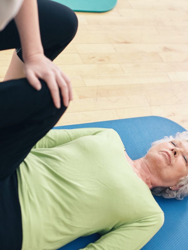 Senior woman lying on exercise mat with her personal trainer helping her doing leg stretches at gym. Elder woman receiving physical training from her personal trainer at rehab.